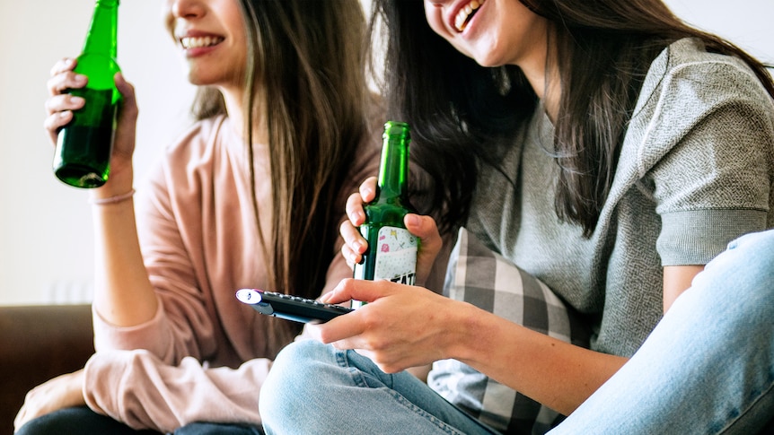 Close-up of two young women with brown hair laughing and drinking beer on the couch.