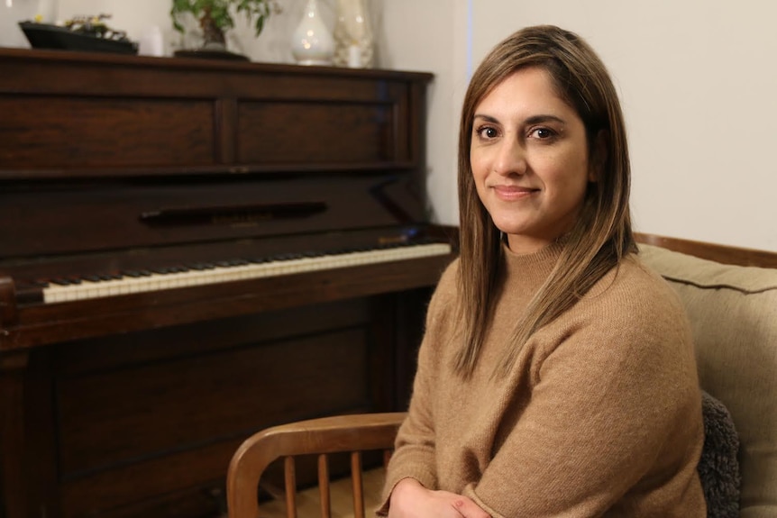 A woman with dark brown hair smiles while sitting in a living room. A stand-up piano is behind her.