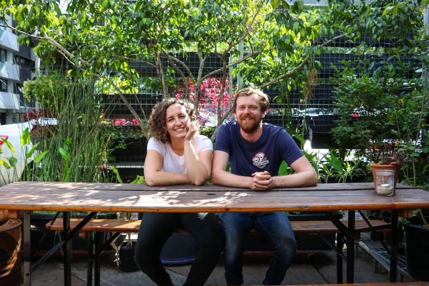 A man and woman sit at a rustic wooden table, smiling, with plants behind them.