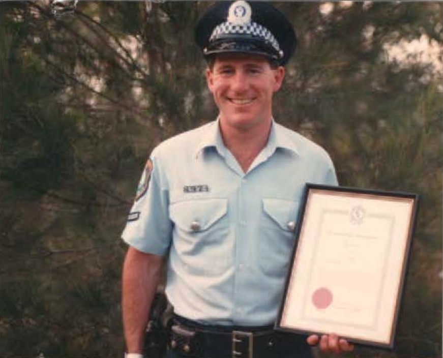 Um jovem Vince Hurley com uniforme de policial sorri e segura um certificado emoldurado.