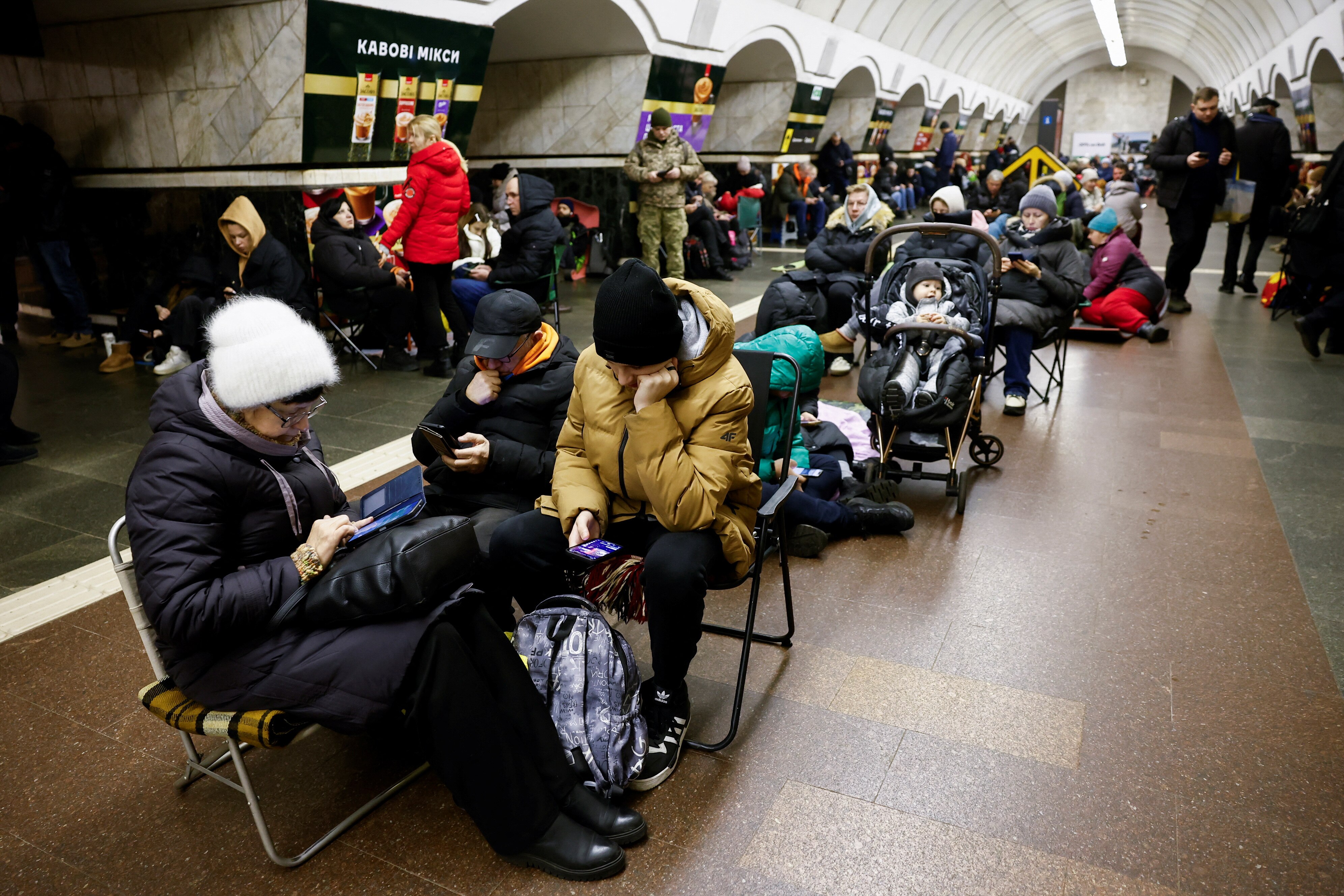 People waring winter coats sitting on chairs and the floor in a line in a hallway of an underground railway station