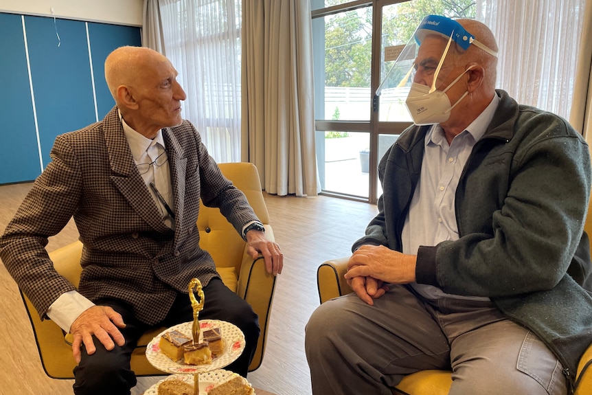 Elderly man sitting next to another man who is wearing a face mask and shield.