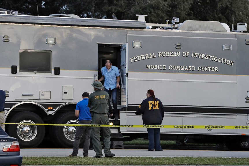 A man exits a grey FBI vehicle with four officials standing next to it and "caution do not enter" tape barring civilians.