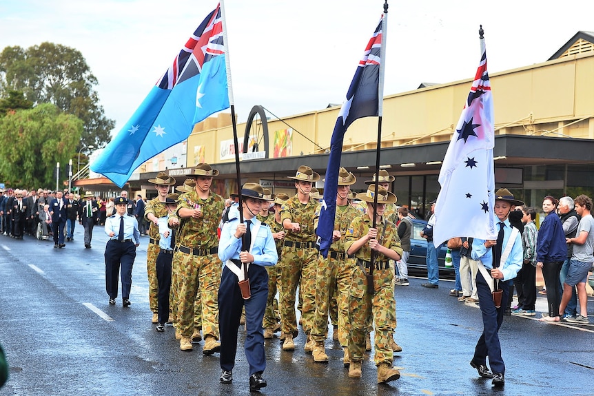 A group of Army cadets march with veterans out the front of a row of shops.