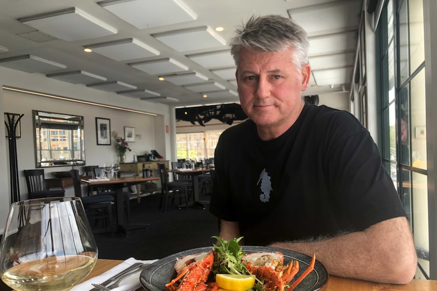 A man sitting at a restaurant table with a plate of lobster and glass of white wine in front of him.