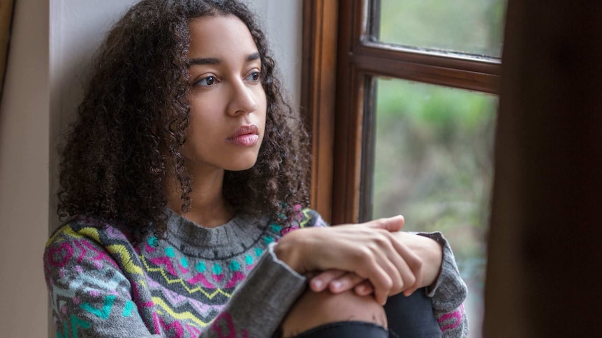 A sad mixed-race teenager sits on the floor against a wall and looks out a window.