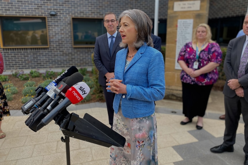 A woman in a blue blazer stands behind a microphone stand, in front of several other at a press conference.