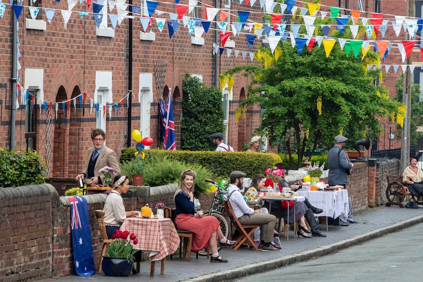 People dressed in 1940s era clothing sit on deck chairs on the footpath in front of their homes