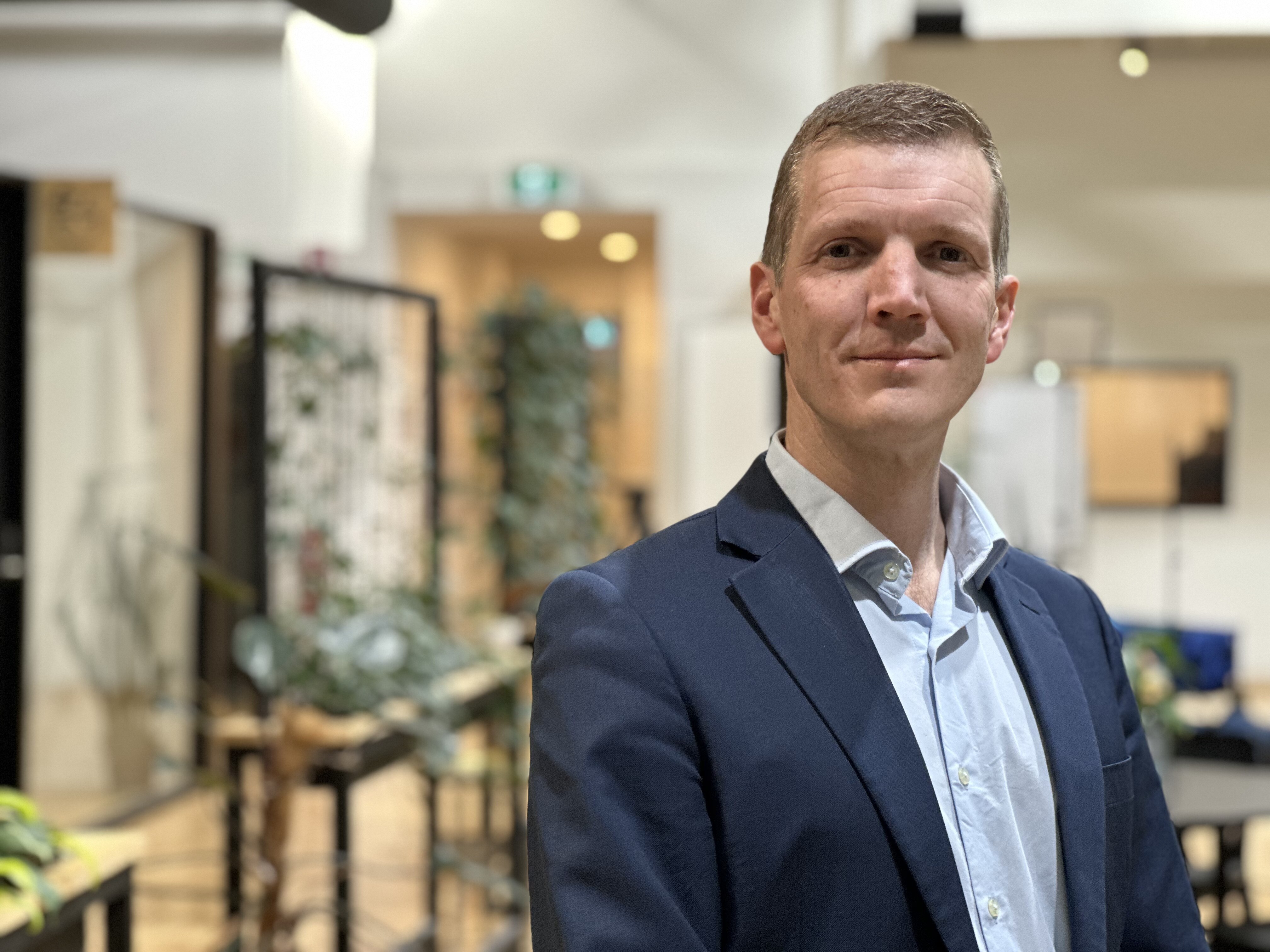 Ross De Rango smiles, standing in a blue suit indoors.