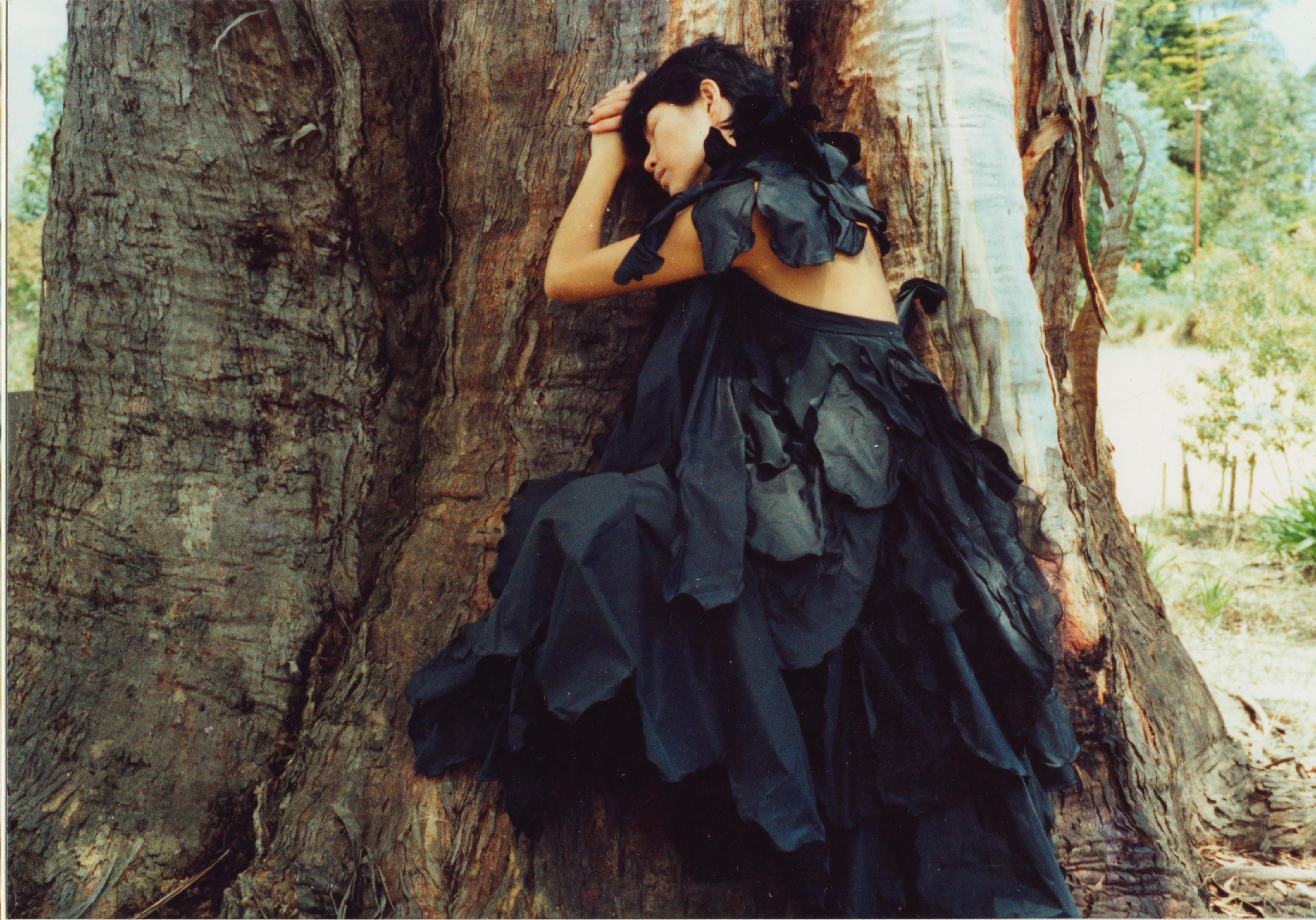 Woman wearing black gown with draping sections like feathers lying against a tree.