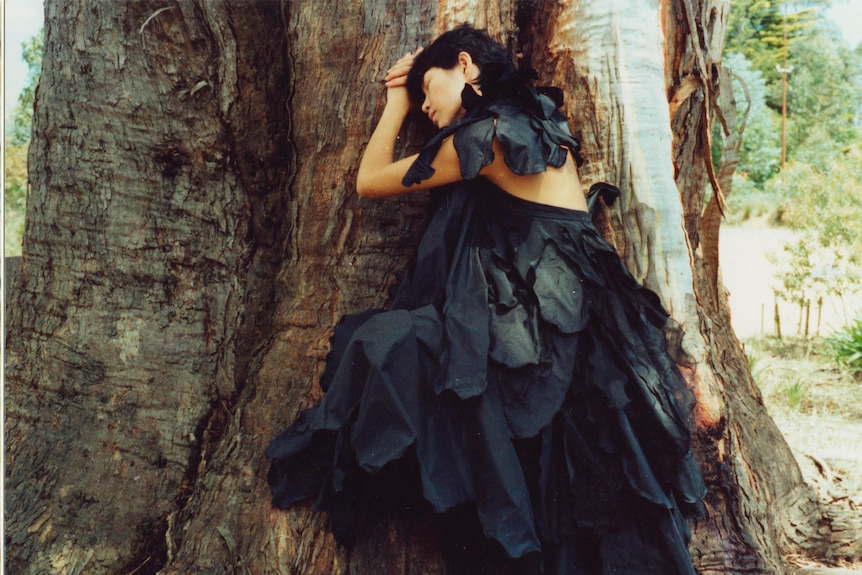 Woman wearing black gown with draping sections like feathers lying against a tree.