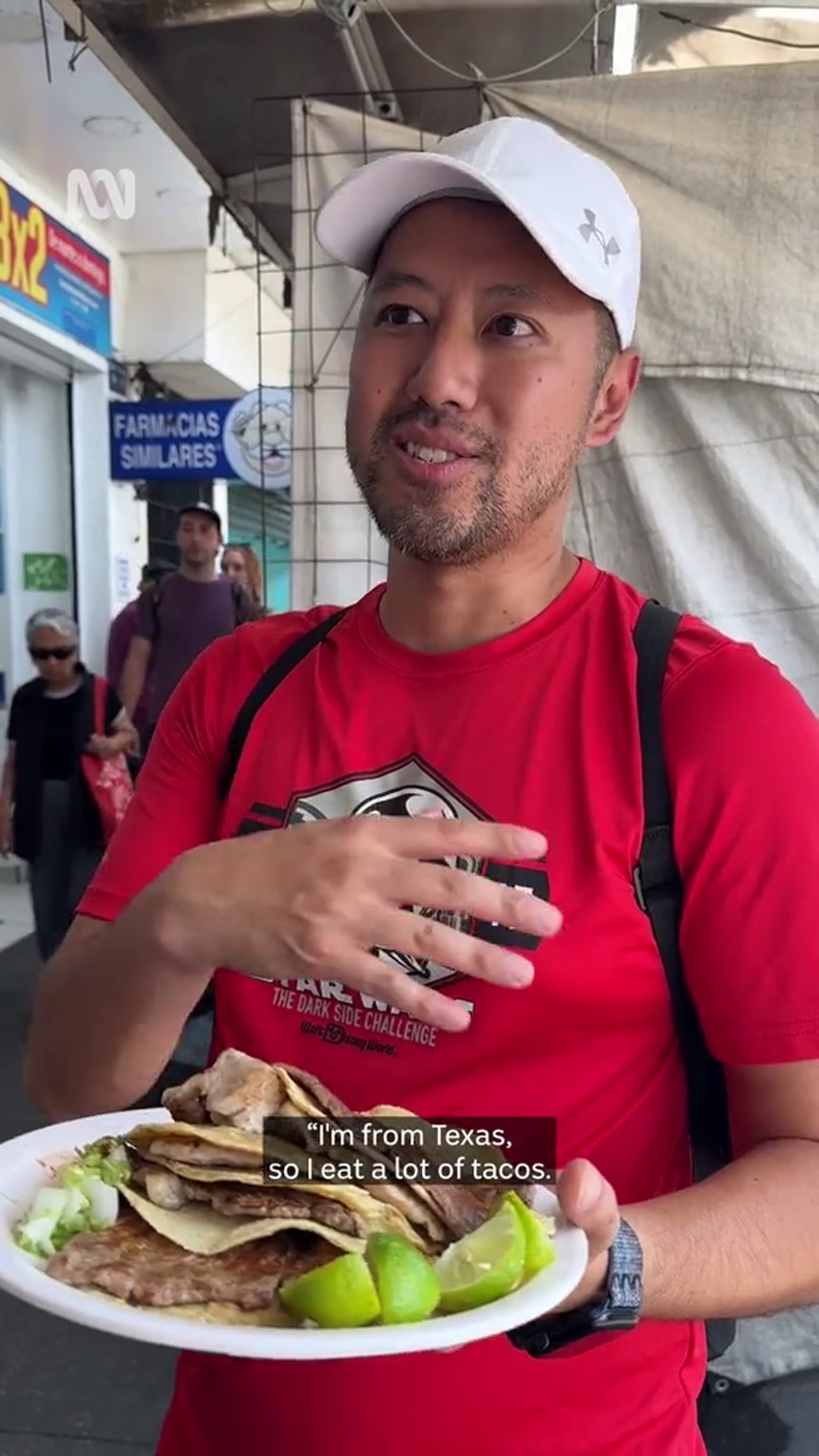 A man with medium-tone skin, dressed in a white cap and red T-shirt holds a plat with tacos and lime on it