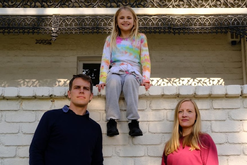 A man and a woman standing in front of a white brick wall with a young girl sitting on top of the wall.