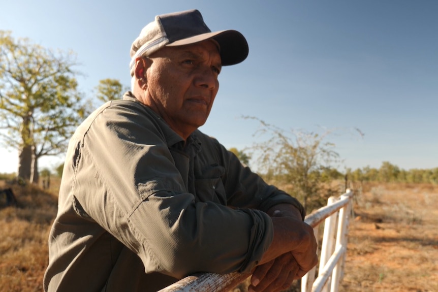A man wearing a cap leans against a fence.
