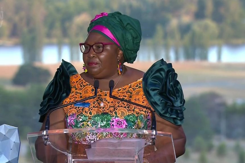 A woman in bright clothing at a lectern makes an acceptance speech