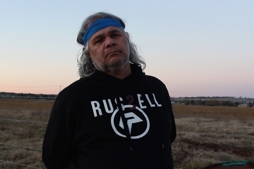 An Indigenous man in a bandana and a hoodie stands outdoors in low light.