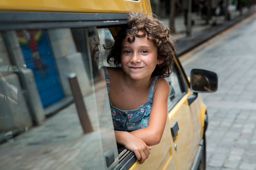 Colour photo from 2017 film Summer 1993 of Laia Artigas learning out the window of a yellow car parked on a cobbled street.