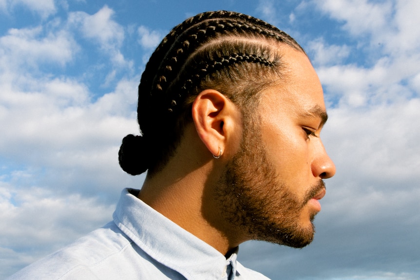A side profile of Ziggy Ramo with cornrows, beard and moustache, blue shirt, blue sky with clouds behind him