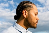 A side profile of Ziggy Ramo with cornrows, beard and moustache, blue shirt, blue sky with clouds behind him