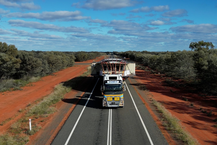 A yellow truck drives through red dust country, hauling an old boat on its trailer and taking up two lanes.