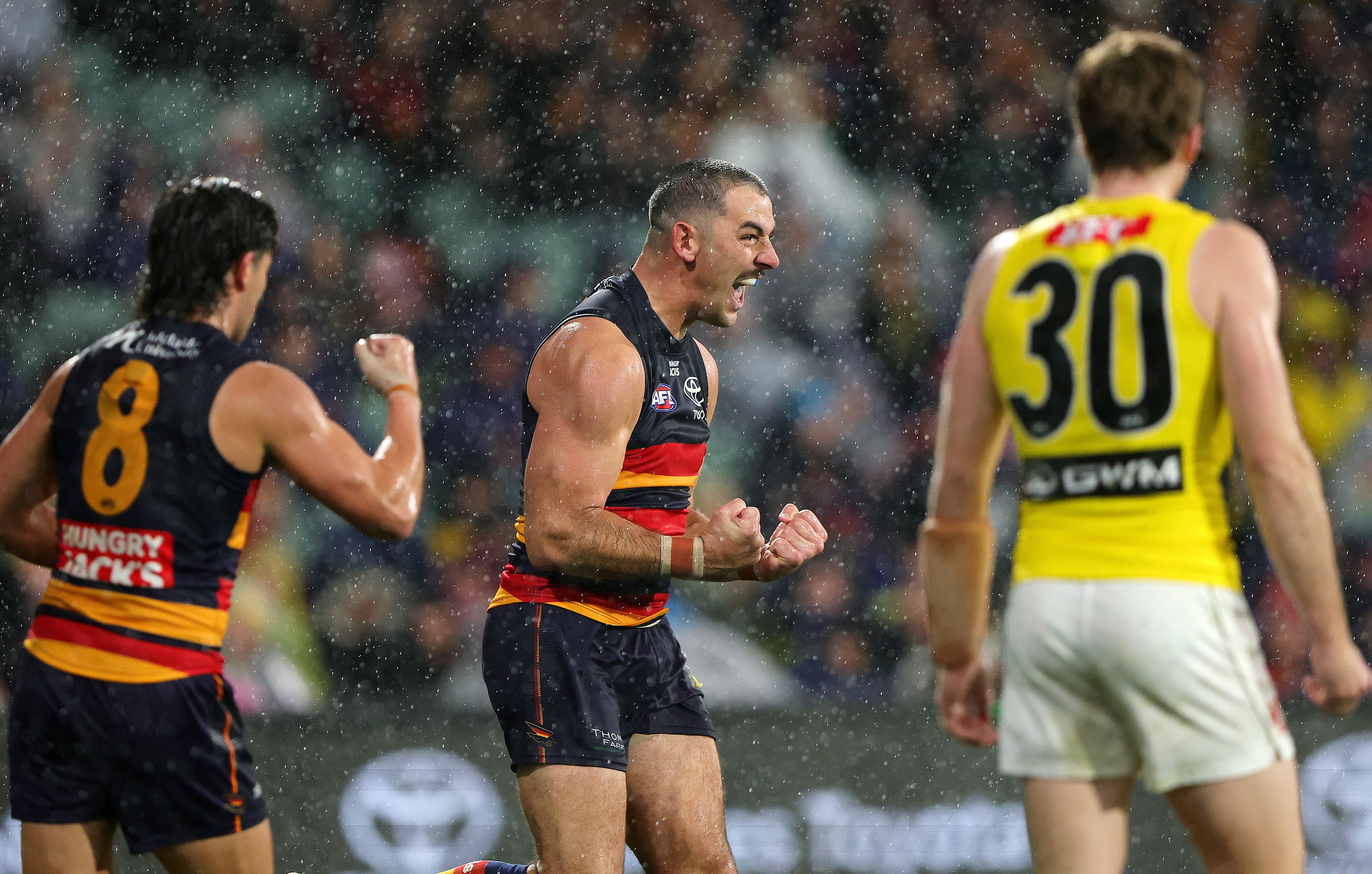 An Adelaide Crows AFL forward roars and pumps his fists as the rain tumbles down after he kicks a goal.