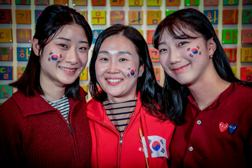 Three Korean women with Korean flags on their cheek, look at the camera and smile.