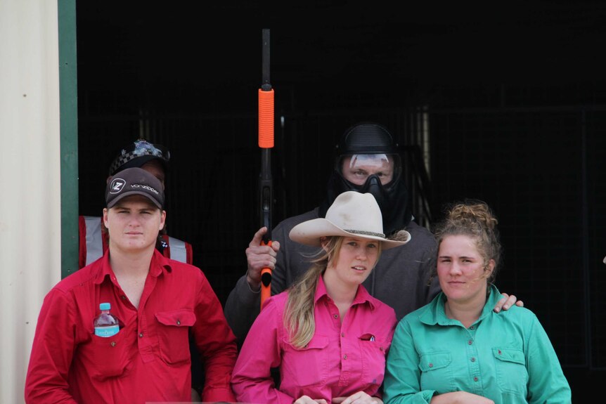 A man in a mask holds a gun and stands with three young people, including one in a cowboy hat.