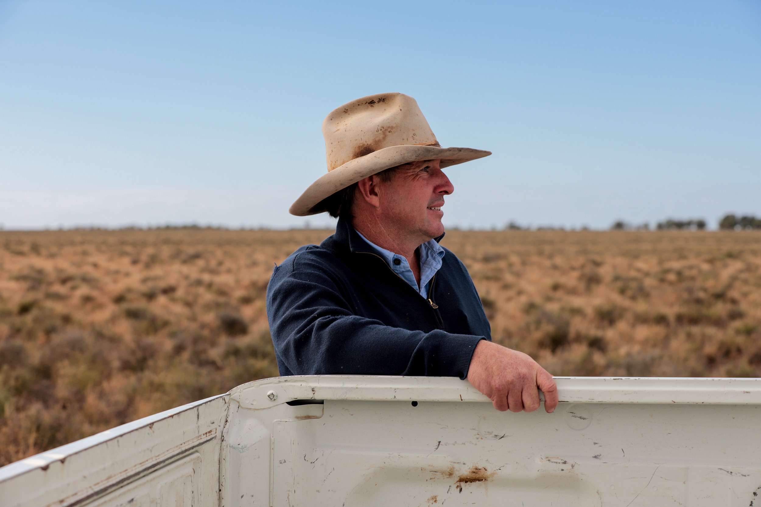 Man wearing weathered wide brim hat has hand on side of white ute tray with flat rural plains behind him