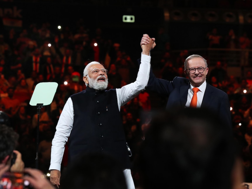 Anthony Albanese holds up Narendra Modi's hand as they stand on stage in an arena. Albanese is smiling. 