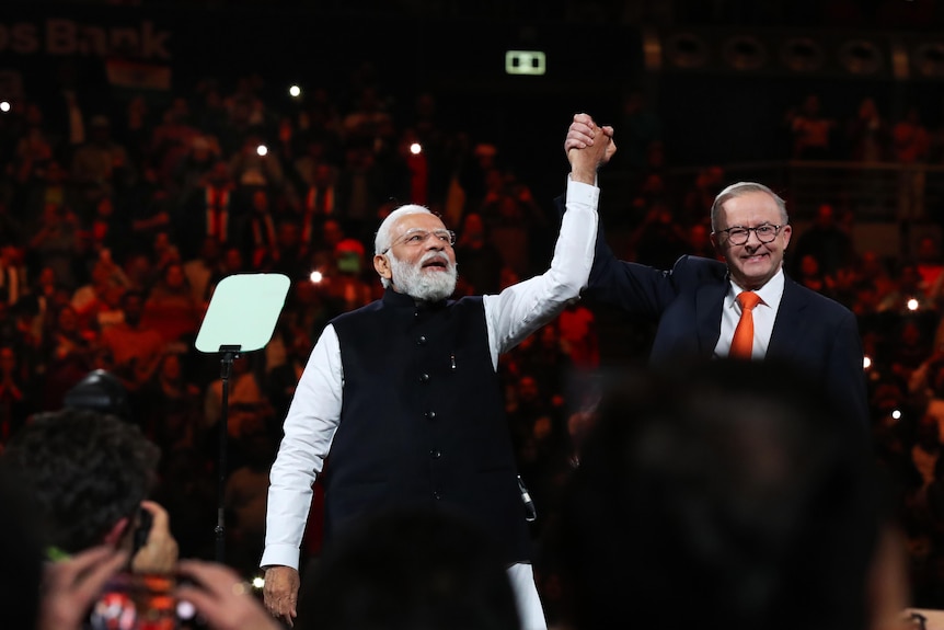 Anthony Albanese holds up Narendra Modi's hand as they stand on stage in an arena. Albanese is smiling. 