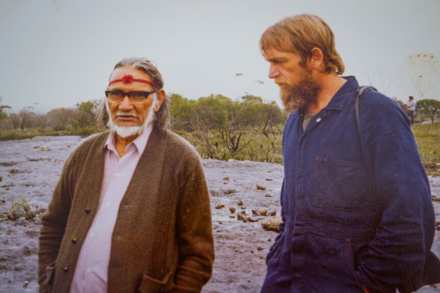 Senior Aboriginal man and younger white man standing together on open ground on a drizzly day.