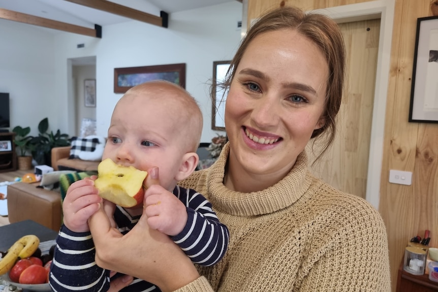A woman wearing a brown jumper holding a toddler and feeding them some apple.