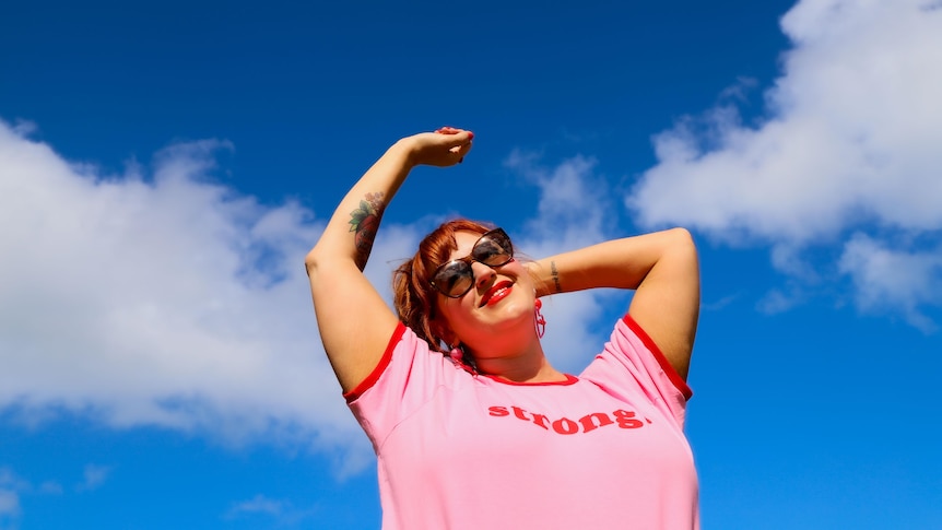 Jen stands in the sunshine, smiling and wearing a pink shirt that visibly reads 'strong'