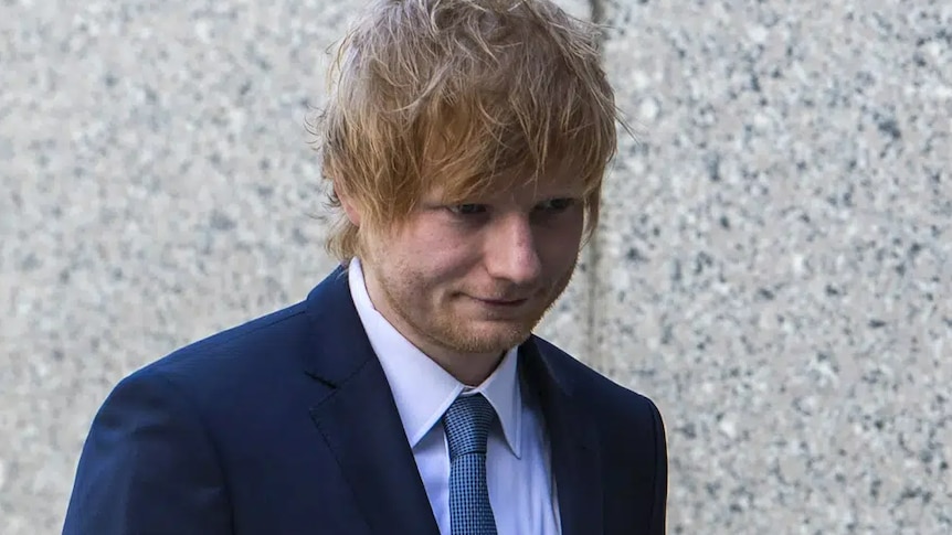 Ed Sheeran wearing a dark blue suit walking past a grey stone wall