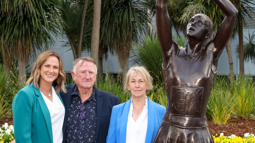 One younger and two older people stand together next to a bronze statue of a young woman shooting a netball