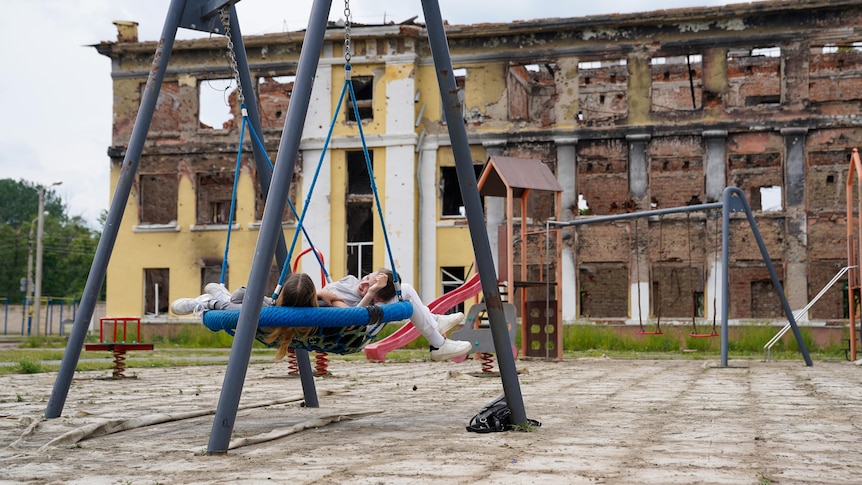 Two young girls recline in a swing chair in a playground. The yellow multi-storey building behind is in ruins