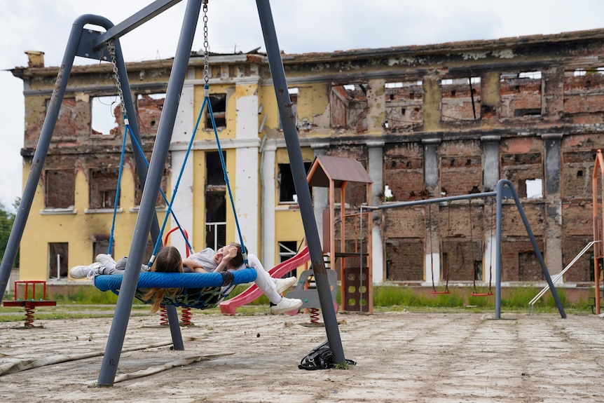 Two young girls recline in a swing chair in a playground. The yellow multi-storey building behind is in ruins