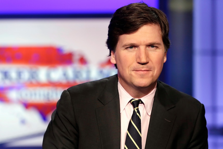 A man with brown hair is wearing a suit and a striped black tie. Behind him is a title screen with text on a map of the US. 