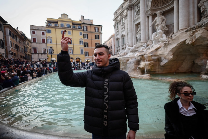 A man in a black puffer coat is ready to throw a coin behind him into the fountain 