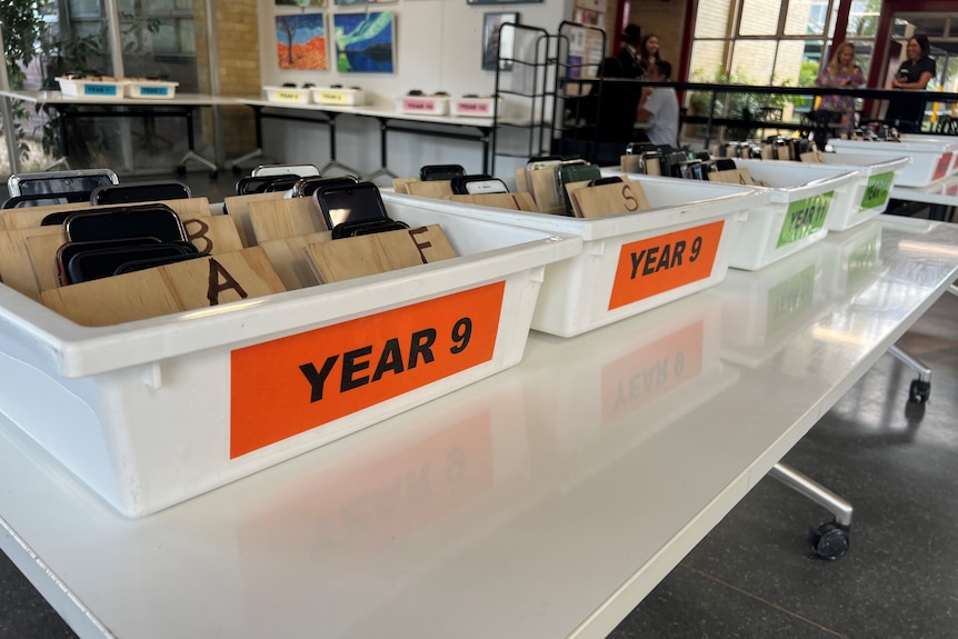 White plastic tubs sitting on a desk with mobile phones inside them and tags such as 'year 9'