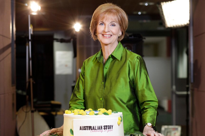 Caroline Jones stands in front of a 20th anniversary cake wearing a green blouse and smiling