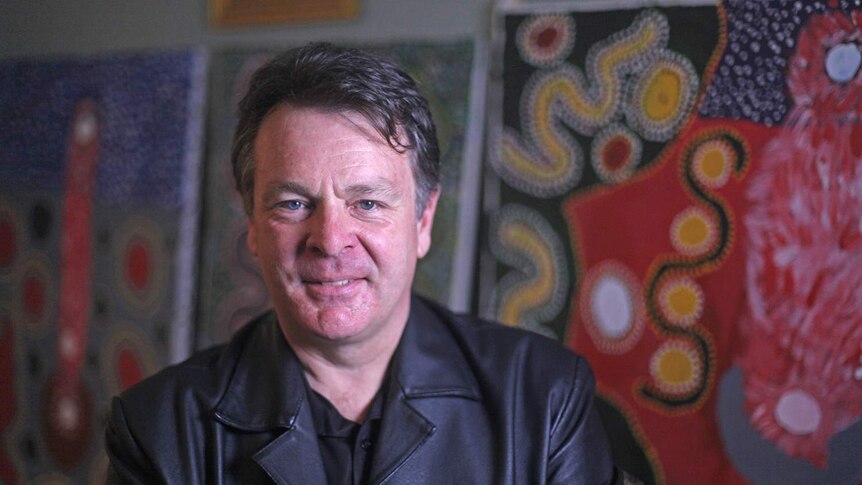 A white man in a back leather jacket sitting in front of Aboriginal artwork.