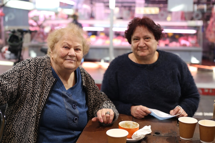 Vicky and her friend smile as they sit at a table with coffees in front of the butcher's section of the market.