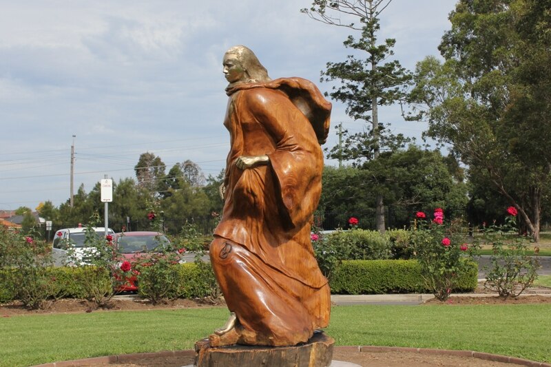 A large wooden sculpture of a woman with flowing robes in front of a rose garden