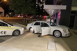 A white sedan with its doors open and tyres flat in front of a police car