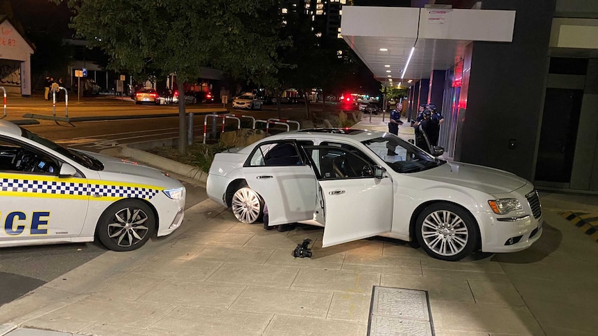 A white sedan with its doors open and tyres flat in front of a police car