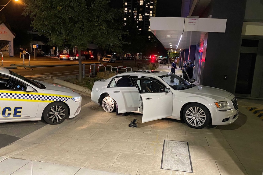 A white sedan with its doors open and tyres flat in front of a police car