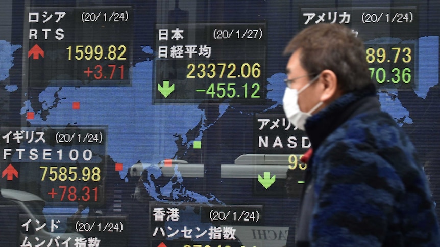 A man in a face mask walks past a stock market sign.