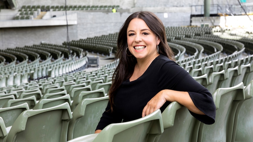 A smiling woman with long dark hair, wearing a dark top, sitting on a seat in a empty grandstand.