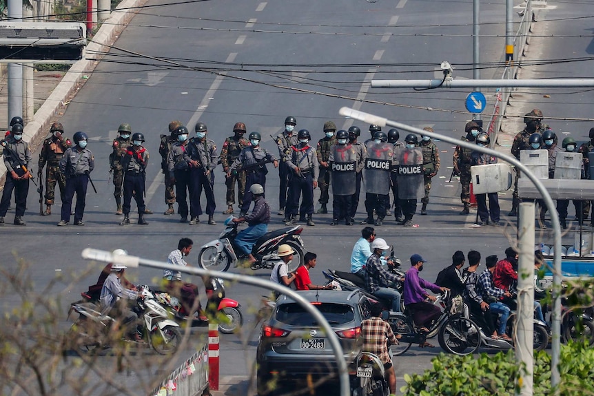 Armed police stand in a line across a main road blocking access while people walk and ride scooters past.